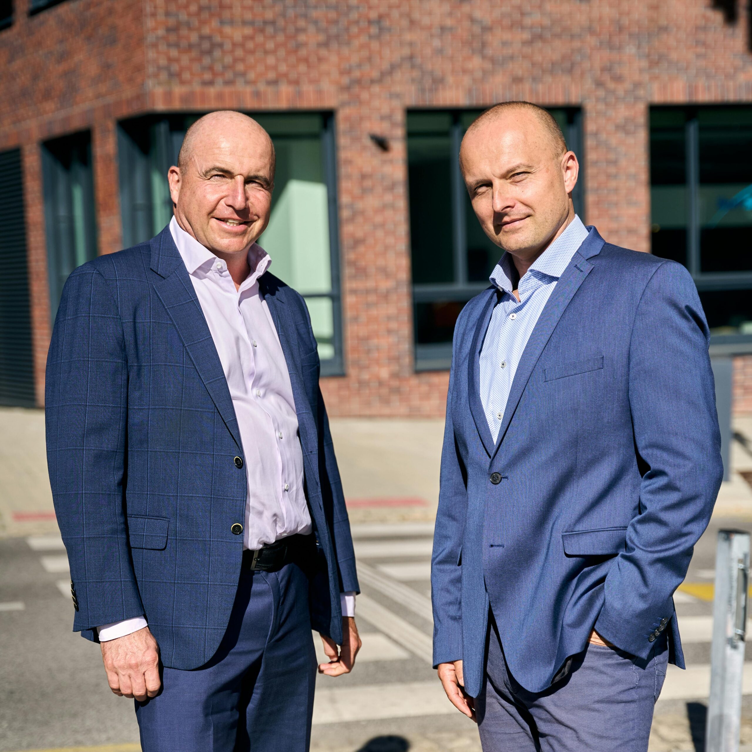 Two businessmen in suits pose confidently outside a brick office building in Bratislava.